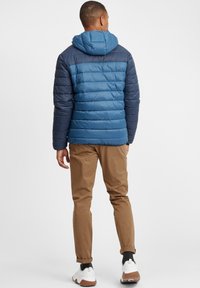 Blå puffjakke med hette, med to-tonet design og horisontale sømmer. Brukt sammen med brune bukser og hvite sneakers.
