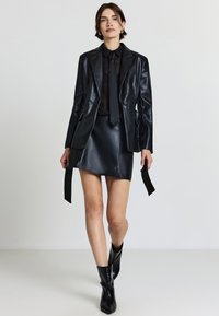 Blazer in pelle sintetica nera con revers a punta, gonna mini coordinata e cravatta nera slim. Una finitura testurizzata e stivali con tacco a punta completano il look.