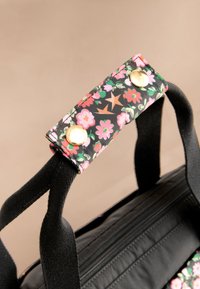 Černá látková taška s květinovým potiskem v růžových a zelených odstínech, zlatým kováním a černými textilními popruhy. Viditelný detail zapínání na zip.