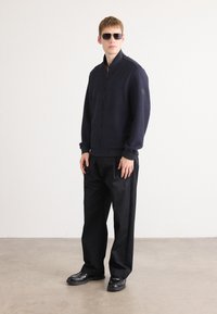 Man in een donker zonnebril, marineblauwe zip-up jas, losse zwarte broek en zwarte schoenen, staande op een betonnen vloer tegen een witte muur.