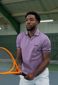 Violet polo marškinėliai su juodais akcentais ir logotipu, balti šortai, laikydamas oranžinį teniso raketę, esan�čiame uždarame lauke.