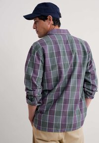 Camisa a cuadros en verde, púrpura y blanco, con una textura suave, mangas enrolladas y una gorra azul marino sólida. Vista desde atrás.