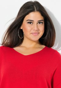 Maglione in maglia rosso a V con maniche corte. Texture morbida, vestibilità rilassata. Grandi orecchini a cerchio in argento mettono in risalto il scollatura.