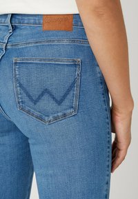 Los jeans de mezclilla azul presentan una etiqueta de cuero marrón en la cintura y un bolsillo trasero con un detalle de costura en forma de "W" característico.