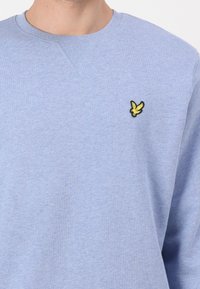 Sudadera azul claro con cuello redondo, puños acanalados y un pequeño logo de un pájaro amarillo bordado en la parte izquierda del pecho. Textura suave.