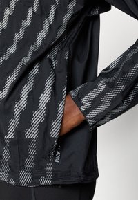 Giacca leggera nera con un motivo a onde riflettenti in argento. Polsini elasticizzati e un'etichetta "PACK IT" sull'orlo.