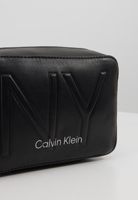 Borsa rettangolare in pelle nera con logo "NY" impresso e testo bianco "Calvin Klein", texture liscia, con dettagli di cucitura superiore.