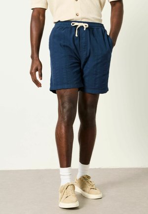 Mann trägt marineblaue Shorts mit Kordelzug, beige kurzärmliges Hemd, weiße Socken und beige Sneaker, steht auf einem grauen Boden.
