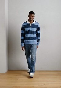 Polo à manches longues rayé bleu et marine avec col, associé à un jean bleu clair et des baskets blanches, se tenant sur un sol en bois.