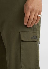 Olive zelene cargo hlače iz gladkega blaga, s preklopno žepom in vezenim logotipom. Roka počiva na žepu, kar poudarja teksturo in obliko.