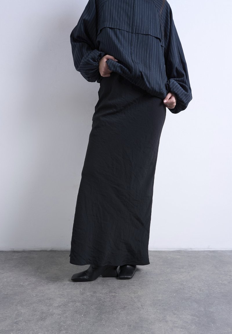 Άτομο που φοράει μακριά μαύρη φούστα, μαύρα μποτάκια με τετράγωνη μύτη και υπερμεγέθη σκούρο μπλε μπλουζάκι με ριγέ, στέκεται σε γκρι δάπεδο μπροστά από λευκό τοίχο.