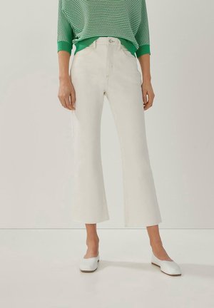 Vrouw draagt witte wijde cropped broek, witte platte schoenen en een groene trui met patroon en driekwart mouwen.