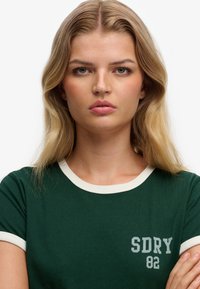 Groen T-shirt met ronde hals en witte geribde kraag en mouwen. Heeft een grijze "SDRY 82" bedrukking aan de linkerkant. Zacht katoenen materiaal.