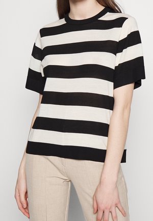 Osoba ubrana w czarno-białą koszulkę z szerokimi poziomymi paskami i krótkim rękawem oraz jasnobeżowe spodnie, stojąca na jednolitym tle.