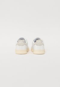 Hvide lædersneakers med en tekstureret beige hælflap, lys cremefarvet sål og 'Stan Smith' logo på bagsiden. Rent, minimalistisk design.