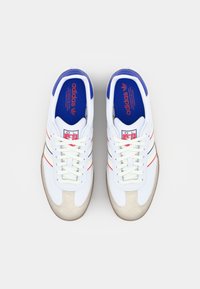 Baskets de sport blanches avec des accents bleus ; dotées de rayures rouges et bleues, d'un capuchon de orteil texturé en beige, et d'un laçage classique. Marque sur la languette.