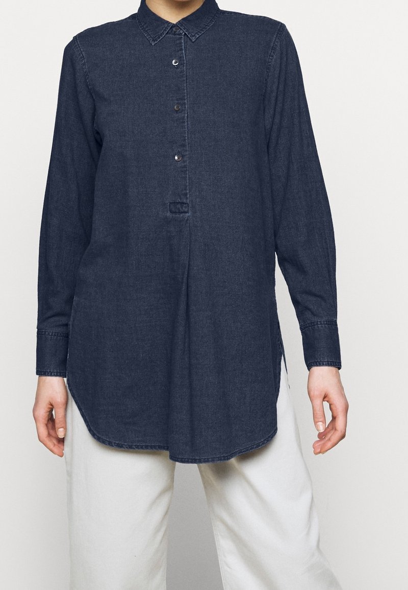 Camicia di denim blu scuro con colletto, apertura con bottoni, maniche lunghe e spacchi laterali; design minimalista con texture liscia.