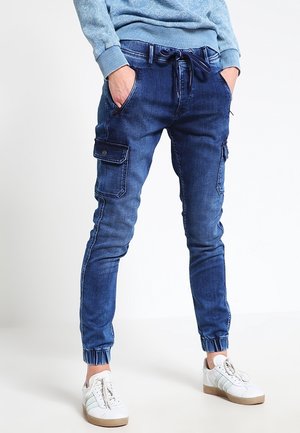 Persoon die blauwe cargo-stijl jogger jeans draagt met elastische manchetten en witte sneakers, handen in de zakken, staand tegen een effen achtergrond.