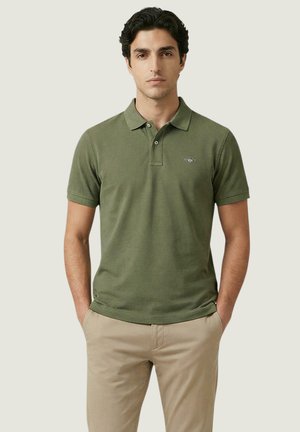 Mann steht mit dem Gesicht nach vorne, trägt ein olivgrünes Poloshirt und beige Hosen, die Hände in den Taschen, vor einem einfarbigen Hintergrund.