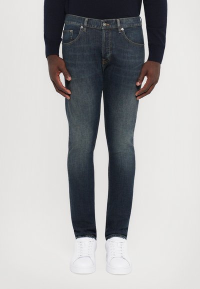 Jeans blu scuro a vestibilità slim, realizzati in denim, dotati di cinque tasche e chiusura con bottoni, abbinati a sneakers bianche.