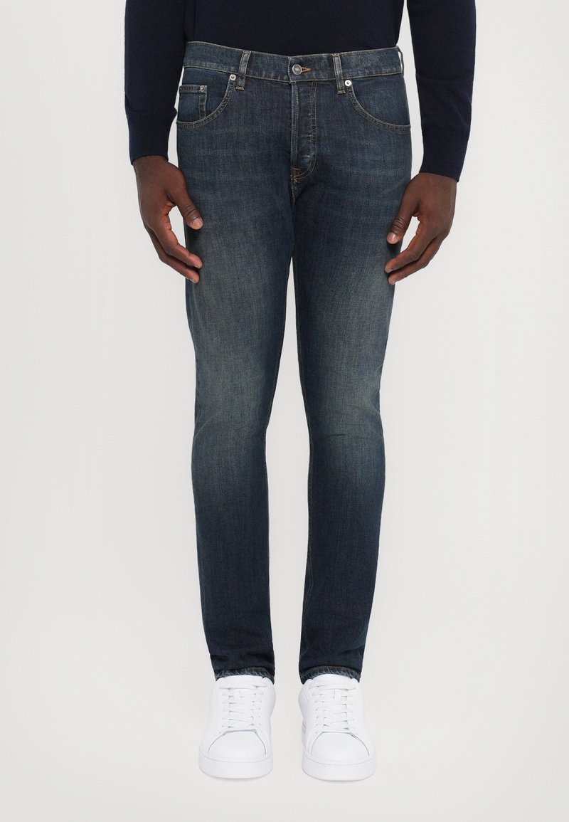 Jeans blu scuro a vestibilità slim, realizzati in denim, dotati di cinque tasche e chiusura con bottoni, abbinati a sneakers bianche.