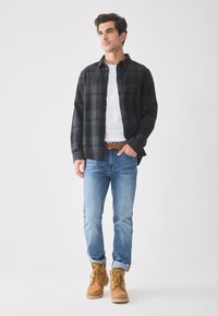 Zwart en grijs geruite flanellen shirt, wit t-shirt, blauwe jeans en tan werklaarzen met gele accenten, allemaal weergegeven op een neutrale achtergrond.