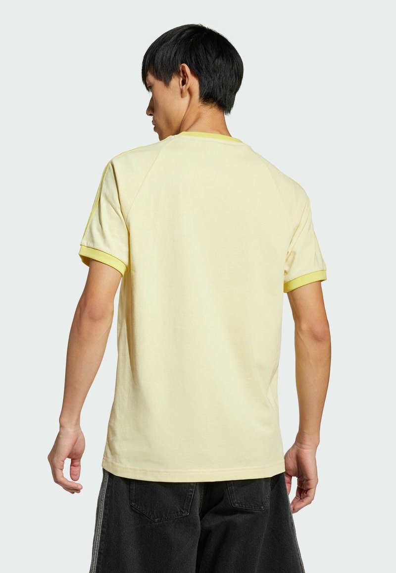 T-shirt di cotone gialla a maniche corte con un bordo di un giallo chiaro a contrasto attorno al colletto e alle maniche; indossata con pantaloni scuri.