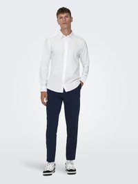 Vit skjorta med lång ärm, parat med marinblå byxor och svarta och vita sneakers. Stående figur med neutral bakgrund.