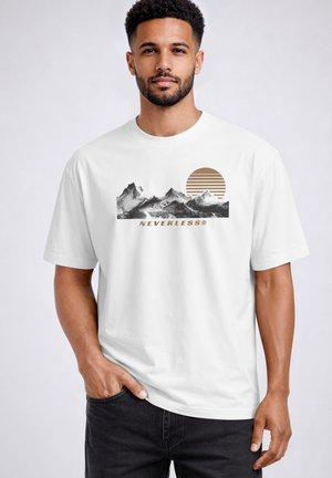 Mann mit kurzen lockigen Haaren und Bart, trägt ein weißes T-Shirt mit schwarzer Berggrafik, braun gestreifter Sonne und dem Schriftzug "NEVERLESS", steht mit der Hand in der Tasche.