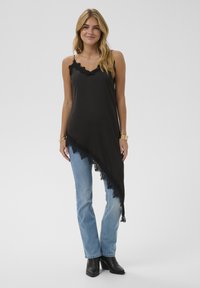 Asymmetrisches schwarzes Camisole mit Spitzenbesatz, getragen über hellblauen Jeans und schwarzen Stiefeletten. Glatte Textur, verstellbare Träger.