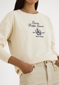 Μπεζ φούτερ με ριμπ μανσέτες, διακοσμημένο με κεντημένο κείμενο και έμβλημα σε ναυτικό μπλε χρώμα. Συνδυασμένο με μπεζ παντελόνι που εμφανίζει λεπτομέρεια πλαϊνής τσέπης.