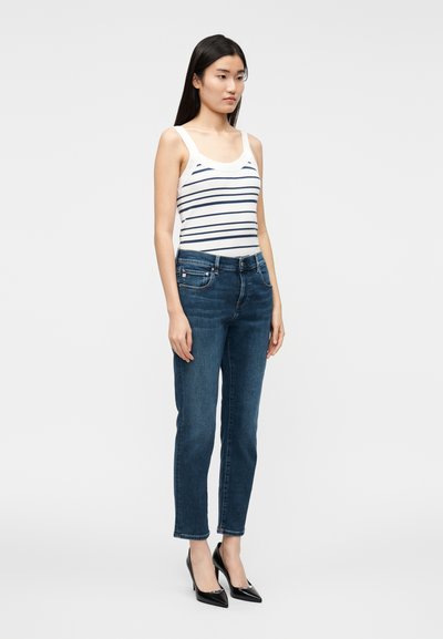 Bijela majica na bretele s tamnoplavim prugama, uparena s tamnoplavim uskim trapericama i crnim cipelama sa špicastim vrhom. Glatki materijal, prianjajući kroj.