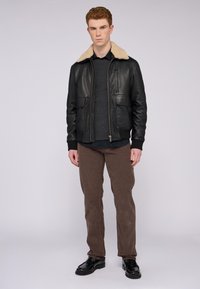 Černá kožená bunda s krémovým kožíškem na límci, dva přední kapsy, nošená přes šedý svetr a tmavé tričko, doplněná hnědými kalhotami.