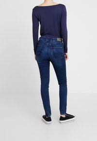 Úzké džíny v tmavě modrém denimovém provedení s hladkou texturou, se zadními kapsami, vysokým pasem a světle hnědou značkovou visačkou viditelnou na pásku.