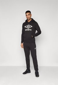 Čierna mikina s kapucňou a bielym logom Umbro, v kombinácii s čiernymi teplákmi s malým logom. Oblečenie má uvoľnený strih a hladkú textúru.