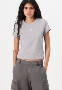 Pilkos spalvos medvilninis marškinėliai su apvaliu kaklu ir trumpomis rankovėmis. Ant krūtinės yra mažas baltas Adidas logotipas. Stilius derinamas su pilkais krovinių kelnais.