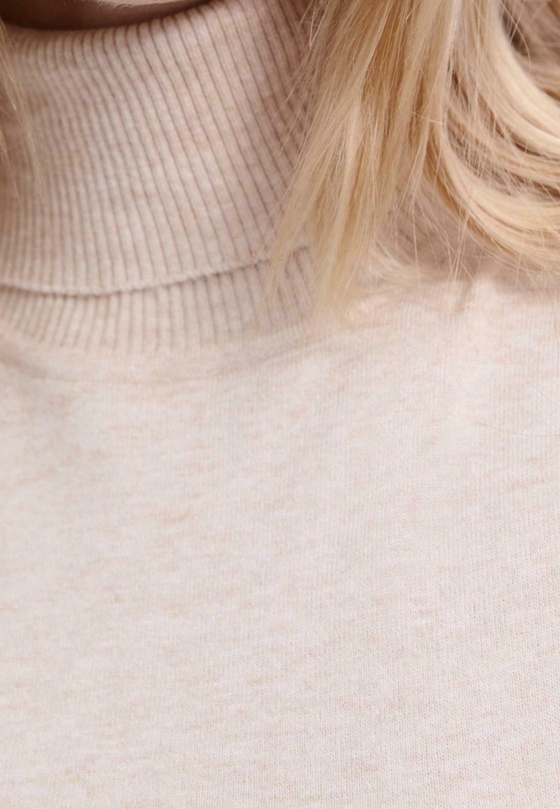 Pull beige côtelé à col roulé porté, avec cheveux blonds mi-longs partiellement visibles.