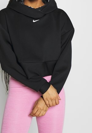 Personne portant un sweat à capuche Nike noir avec le logo sur la poitrine et des leggings rose clair, debout les mains croisées devant.