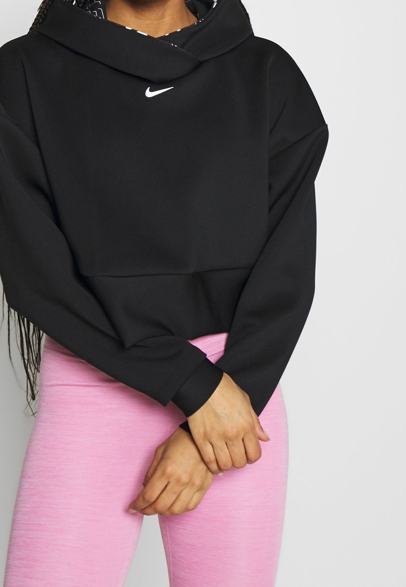Personne portant un sweat à capuche Nike noir avec le logo sur la poitrine et des leggings rose clair, debout les mains croisées devant.