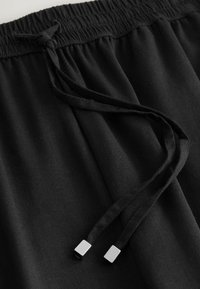 Pantaloni sport negri cu talie elastică adunată și șnur, terminați cu aglete metalice pătrate. Textură moale și fină a țesăturii.