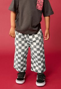 T-shirt gris avec texte imprimé rose, associé à un pantalon large à carreaux noir et blanc et des baskets noires avec des semelles blanches. Fond rouge.