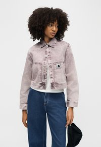 Γυναίκα με σγουρά μαλλιά που φοράει ένα ξεθωριασμένο ροζ κοντό μπουφάν και μπλε τζιν, κρατώντας ένα μαύρο καπέλο στο ένα χέρι.