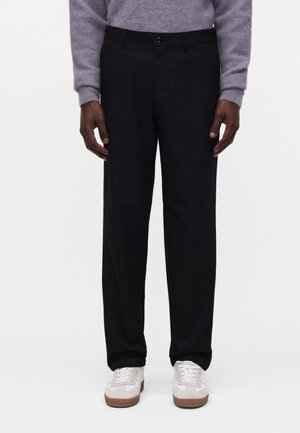 Mann trägt schwarze maßgeschneiderte Hose, grauen Pullover und weiße Turnschuhe, steht vor einem schlichten weißen Hintergrund.