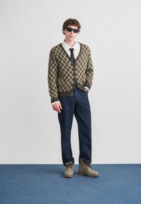 Καρντιγκάν καρό ελιάς και μαύρου χρώματος πάνω από λευκό πουκάμισο και μαύρη γραβάτα, συνδυασμένο με σκούρο μπλε τζιν και μπεζ μπότες σε μπλε χαλί.