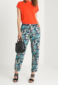 Kobieta ubrana w jasnopomarańczową bluzkę z krótkim rękawem, niebieskie spodnie w kwiatowy wzór, czarne sandały na obcasie i trzymająca małą czarną torebkę.