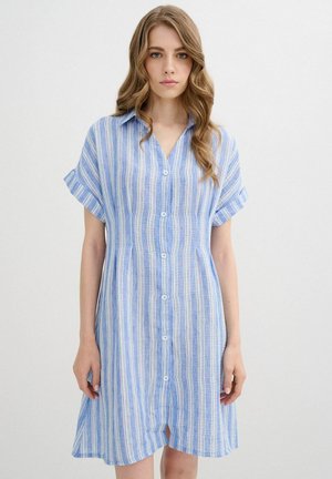 Joven mujer con cabello ondulado que lleva un vestido de rayas azules y blancas con botones en la parte delantera y mangas cortas, posando frente a un fondo liso.