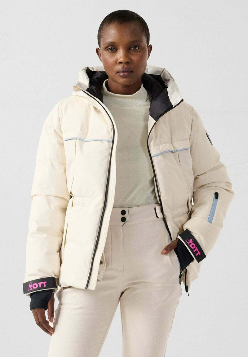 Giacca a piumino color crema con dettagli neri, tasche con cerniera e logo rosa sulle maniche; dotata di cappuccio e texture trapuntata.