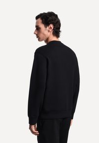 Giovane uomo con capelli corti e scuri che indossa un semplice felpa nera, mostrato di spalle e leggermente girato a sinistra su uno sfondo neutro.