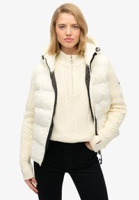 Kurtka puchowa w kremowym kolorze z teksturowanymi rękawami i czarnym zamkiem. Posiada kaptur i wysoki kołnierz, noszona na kremowym swetrze w splot kablowym.