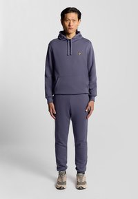 Lila sweatshirt med ficka fram och dragsnodd i huvan, ihopparad med matchande lila joggers. Har en liten gul logotyp på bröstet.